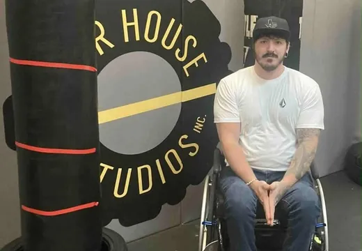 An adaptive athlete in a wheelchair beside the Our House Studios logo wall